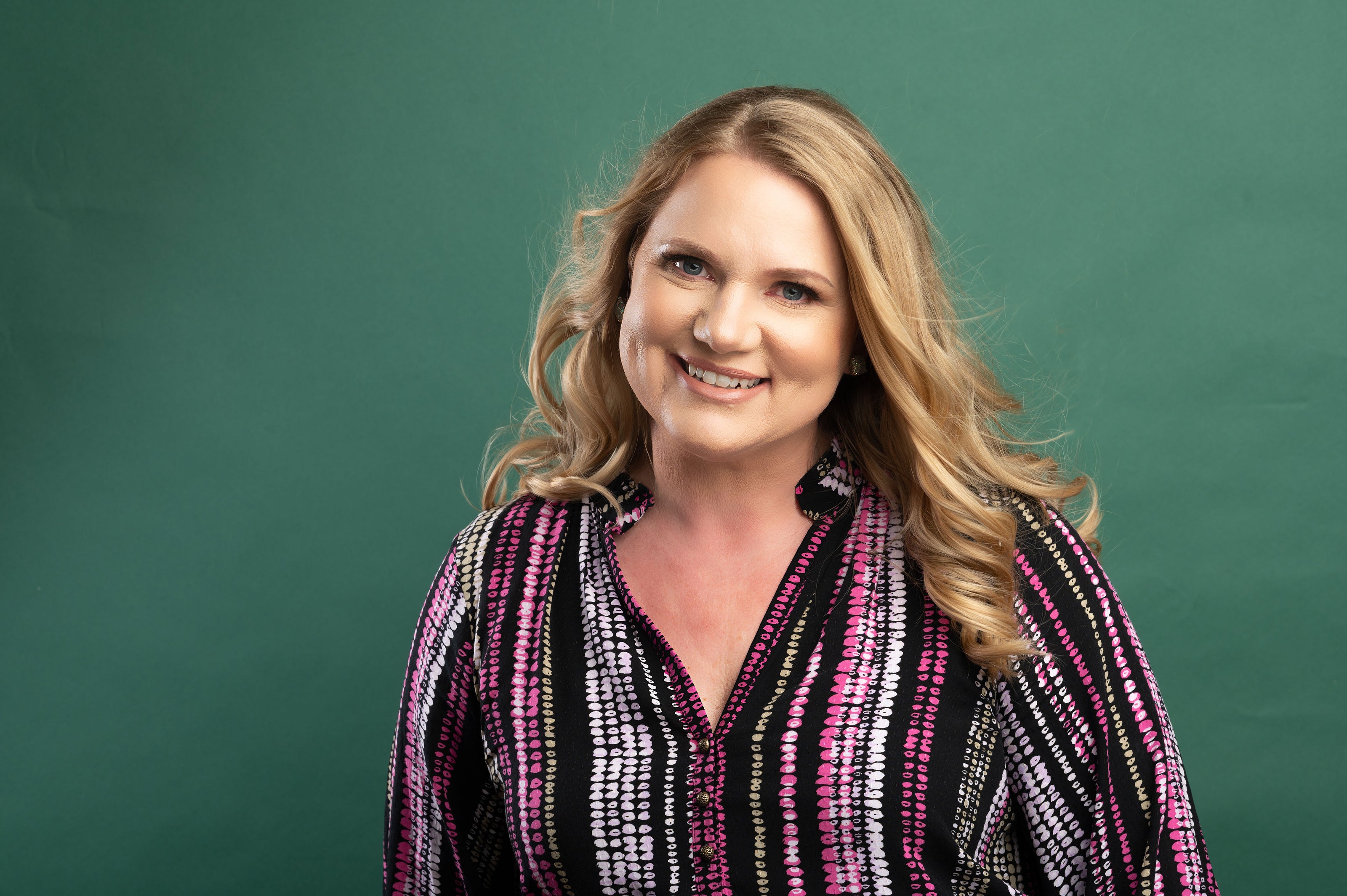 Woman with long blonde hair wearing a patterned shirt against a green background