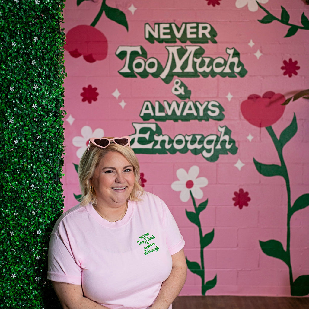 Woman wearing pink tshirt that says "Never Too Much & Always Enough" in small green font near shoulder
