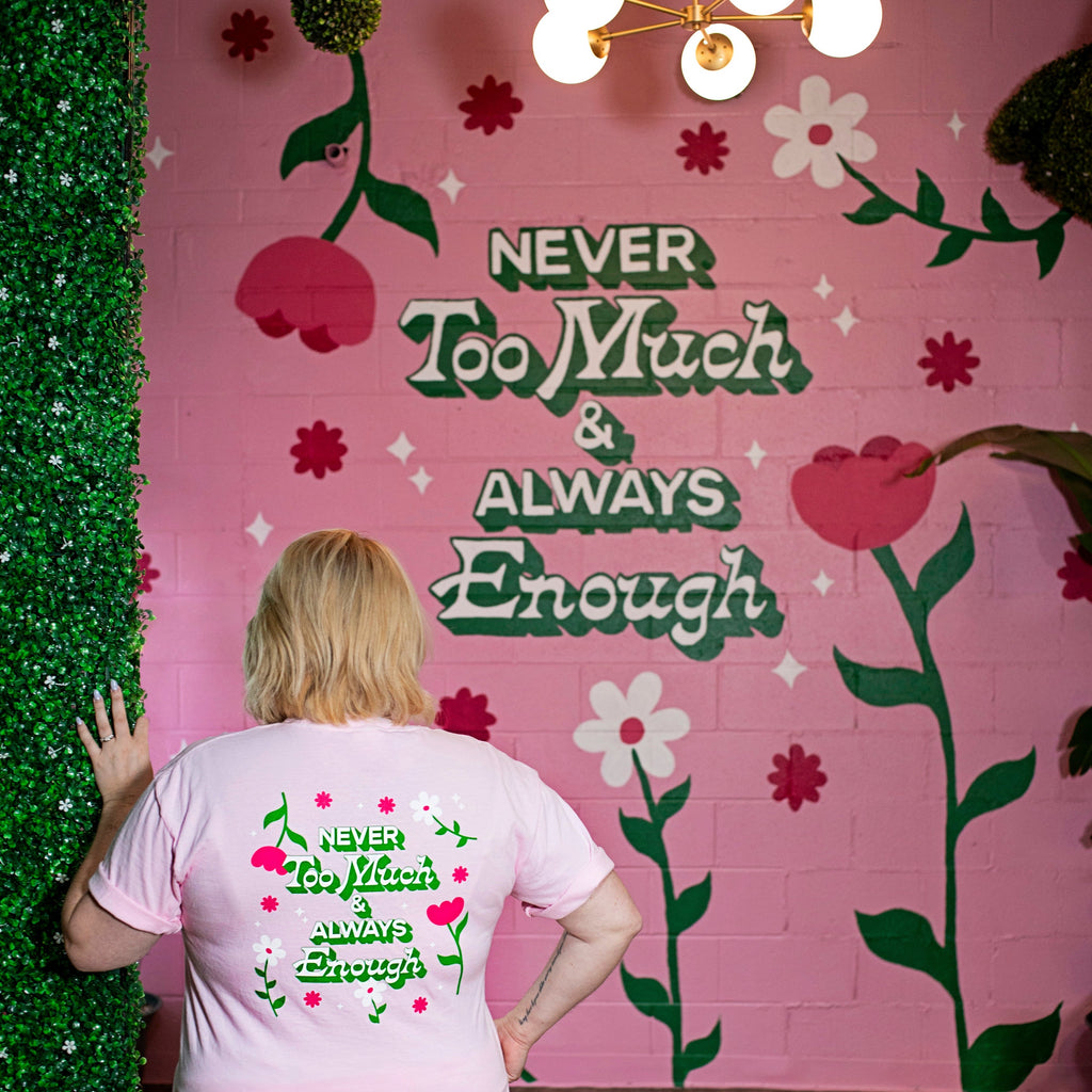 Woman showing off the back view of a tshirt that says "Never Too Much & Always Enough" surrounded by florals 