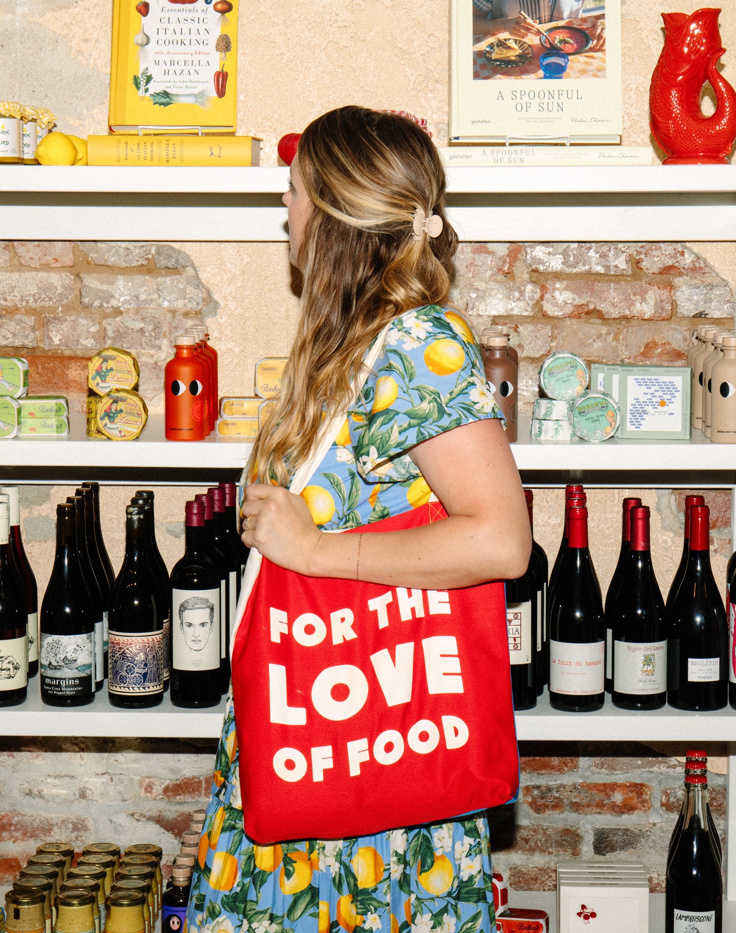 Woman holding a red tote bag with 'For the Love of Food' text in a store setting.