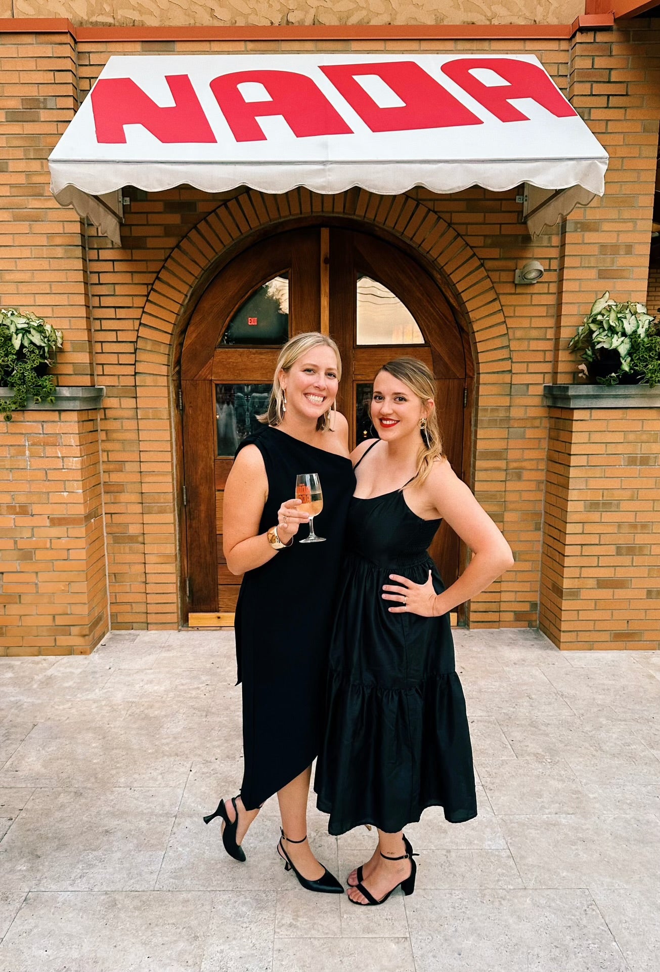 Two women in black dresses standing in front of a building with a 'Nada' sign.