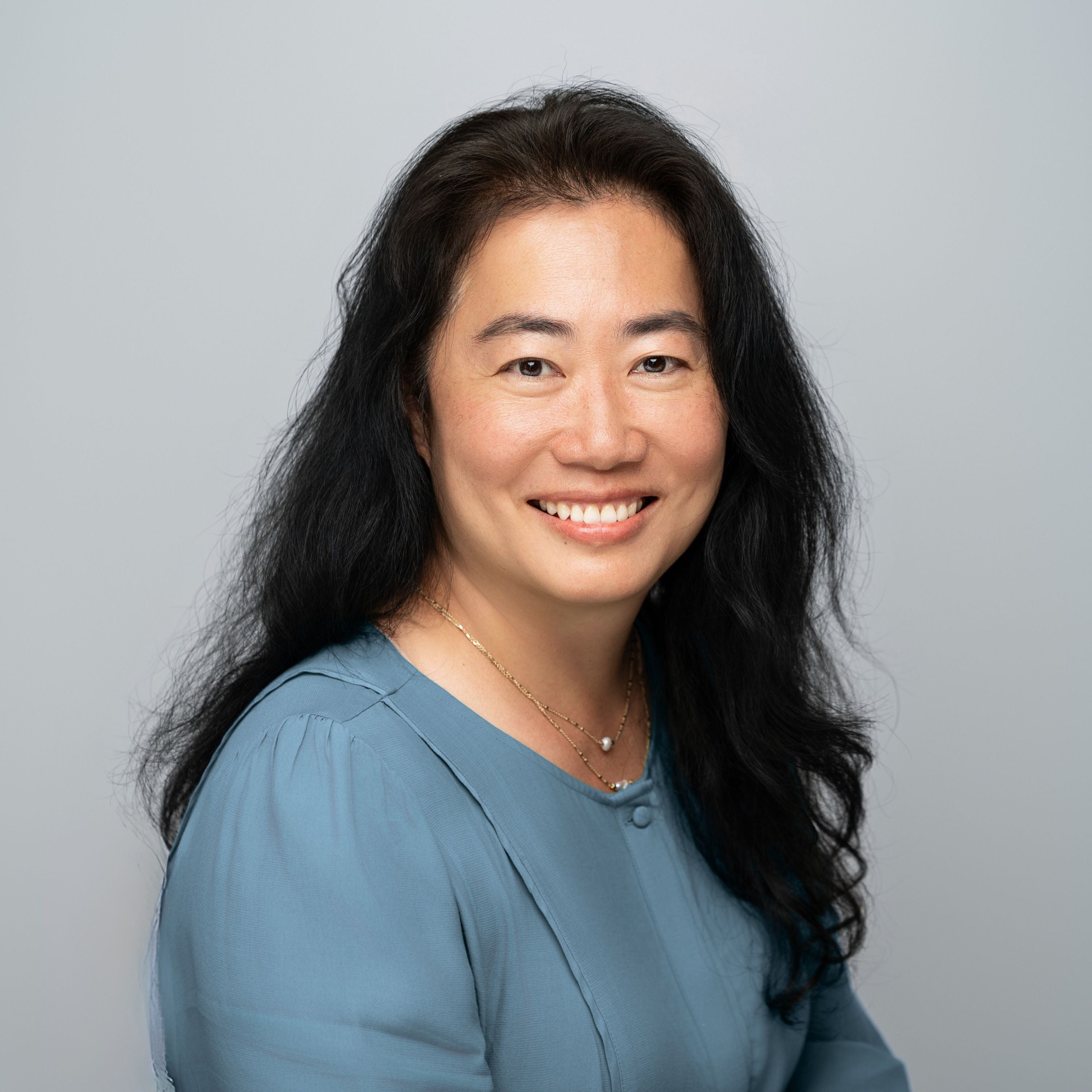 Woman with long dark hair wearing a blue top against a gray background