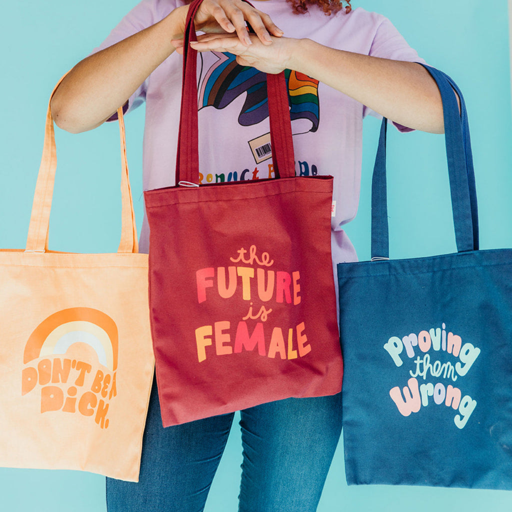 woman holding three totes with the phrases "Don't Be A Dick", "The Future Is Female" and "Proving Them Wrong"