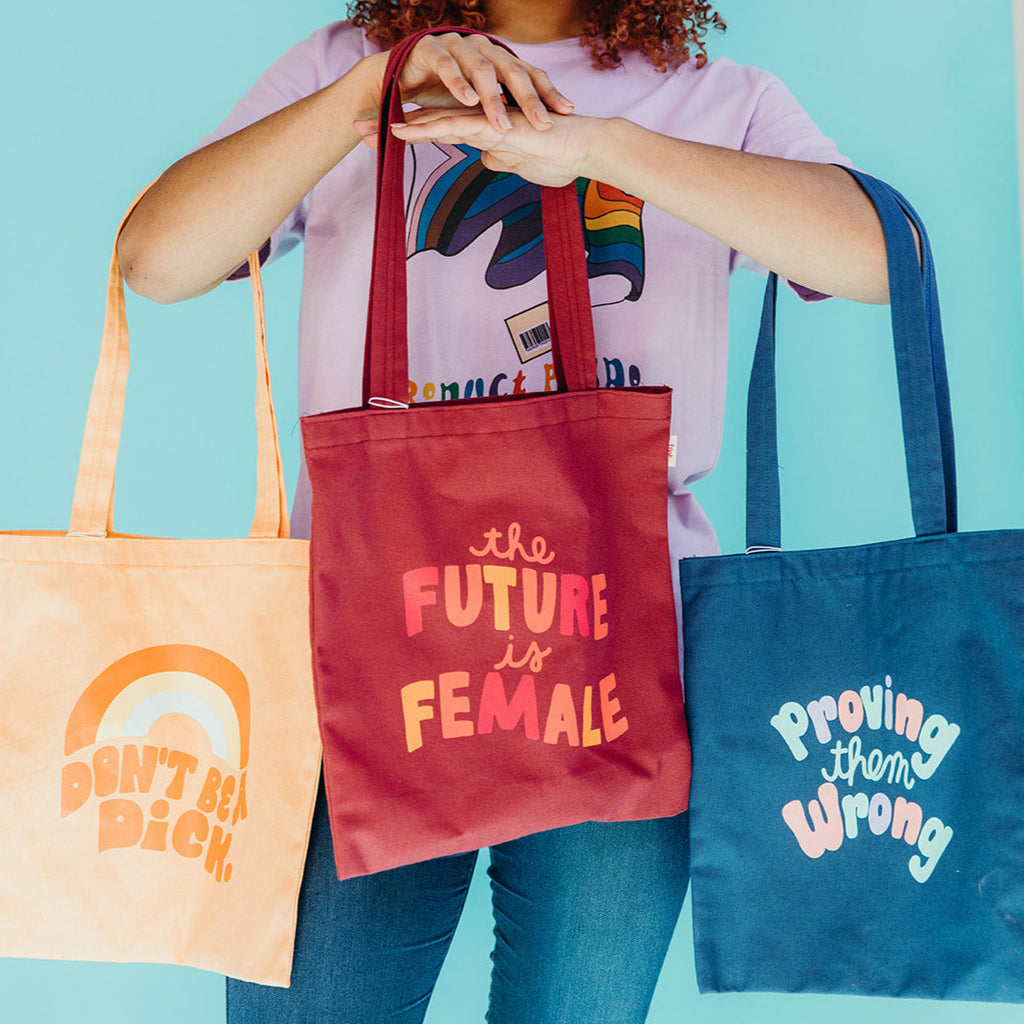 woman holding three totes featuring the phrases "Don't Be A Dick", "The Future Is Female" and "Proving Them Wrong"