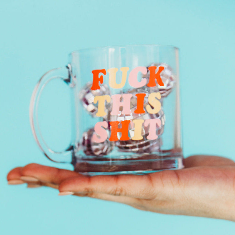 hand holding clear glass Fuck This Shit mug against a turquoise background