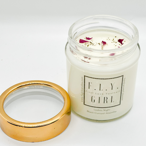 White candle in a clear glass jar with a gold lid laying next to it. The candle has dried florals on top. This candle is from Fly Girl Candles in the scent "Ladies Night".