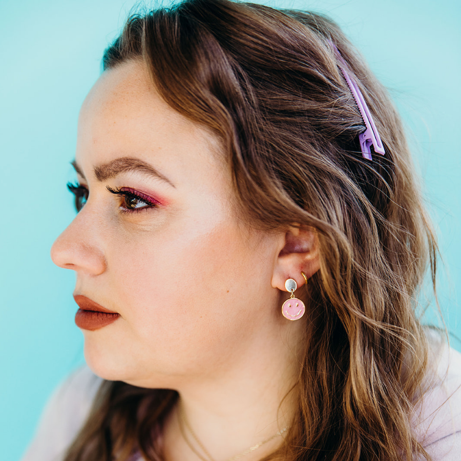 close up of woman wearing the Be Happy smiley face earrings in pink against turquoise background
