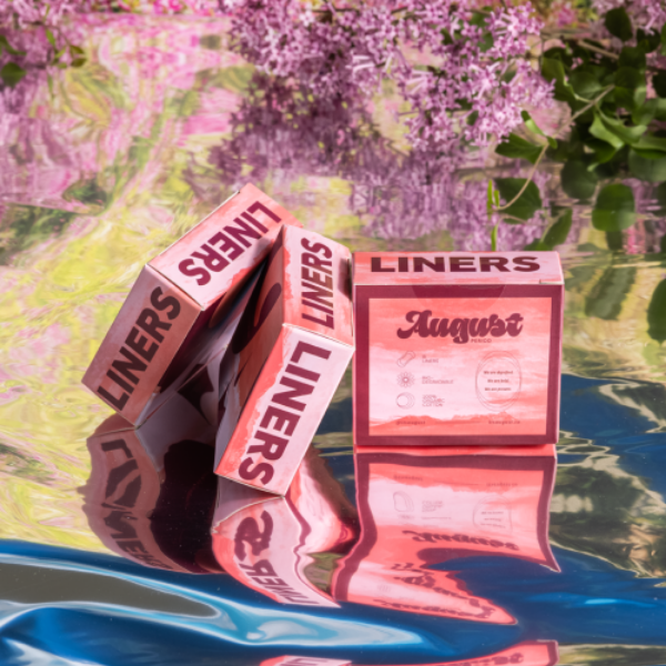 Pink and red boxes of period liners from the brand August laying on a reflective backdrop that resembles a pond surrounded by pink flowers. 
