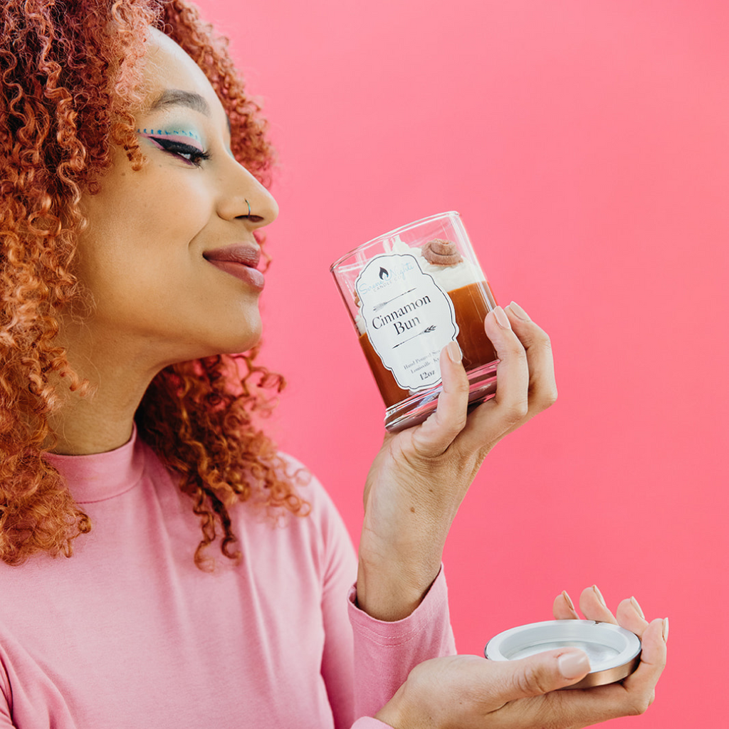 close up of woman smelling Cinnamon Bun candle against pink background