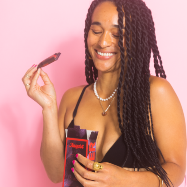 Woman with long black braided hairstyle smiling and looking down at boxes of super tampons from period care brand August. She is pulling one of the tampons of out the box. Woman is standing in front of a soft pink backdrop.