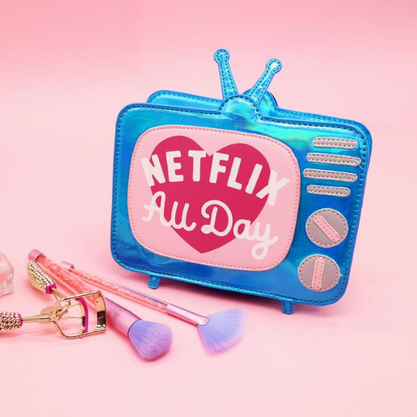 Blue iridescent novelty bag that is shaped like a vintage television set. Features a pink &quot;screen&quot; with a dark pink heart and the words &quot;Netflix all day&quot;. Also features silver dial knobs. The purse is sitting on a pink backdrop with makeup brushes in front of it.