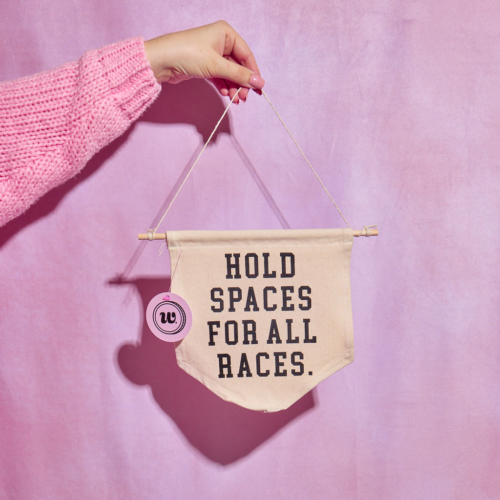 Model holding an ivory wall hanging that says "Hold spaces for all races" in black ink in front of a pink backdrop. Wall hanging sold at Woman-Owned Wallet, a feminist gift shop in Louisville KY.