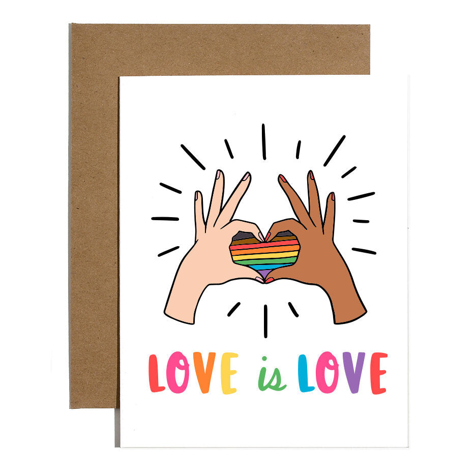 White greeting card with hand drawn illustration of two hands coming together to create the shape of a heart, surrounding the LGBTQIA+ flag. The phrase "love is love" is written underneath the illustration in rainbow colors. There is a brown kraft envelope underneath the card.