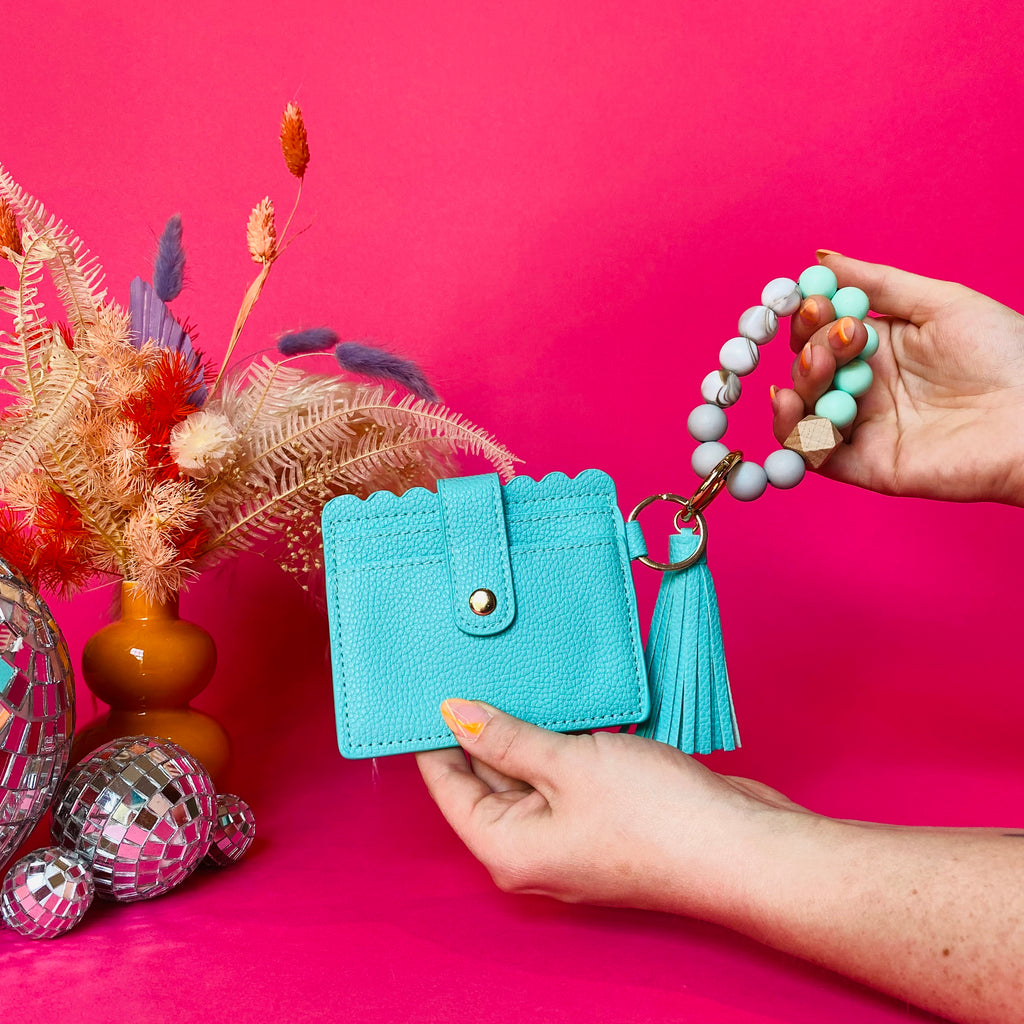 Closeup of woman's hands holding a teal faux leather wallet featuring two back card pockets, a snap closure, attached bracelet made from grey and teal silicone beads, and a purple faux leather tassel. Pink backdrop with an orange vase filled with dried florals sitting next to small disco balls.