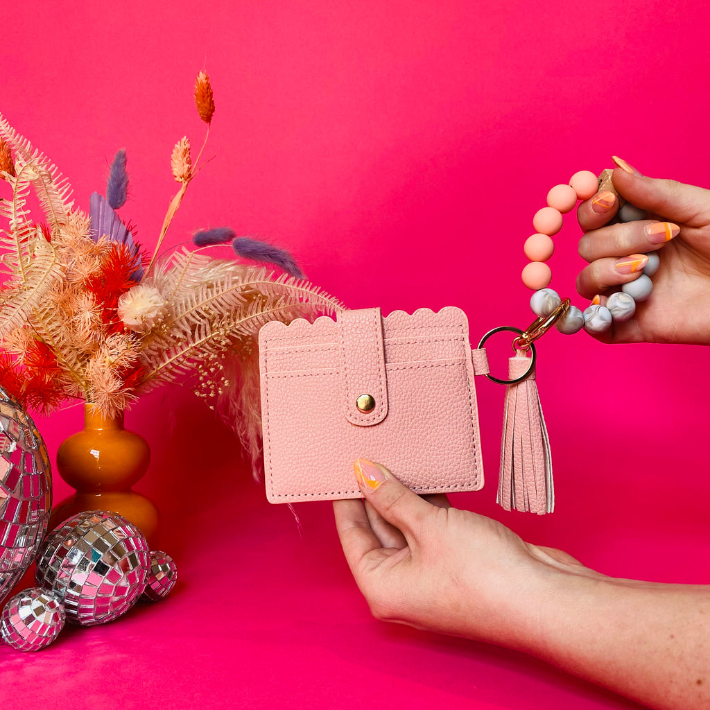 Closeup of woman's hands holding a peach faux leather wallet featuring two back card pockets, a snap closure, attached bracelet made from grey and peach silicone beads, and a peach faux leather tassel. Pink backdrop with an orange vase filled with dried florals sitting next to small disco balls.