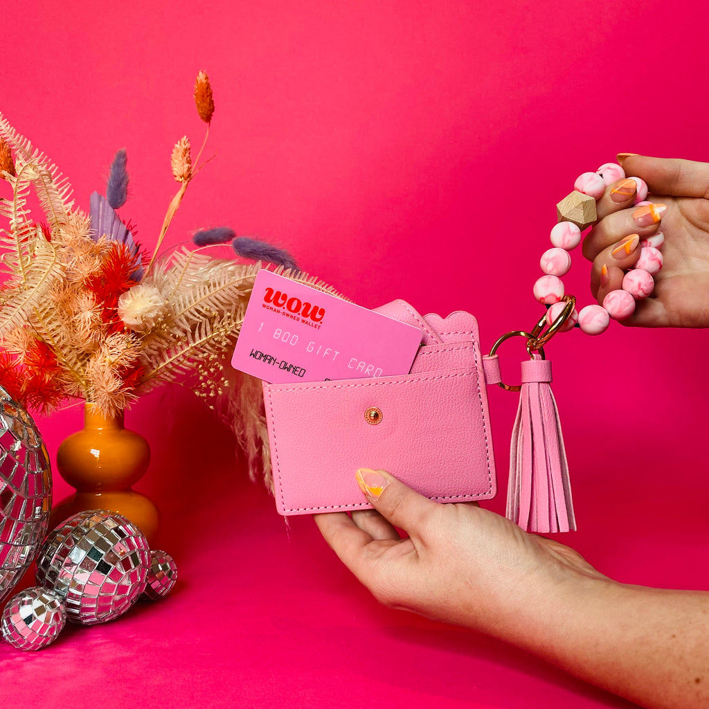 Closeup of woman's hands holding a pink faux leather wallet featuring two back card pockets, a snap closure, attached bracelet made from pink silicone beads, and a pink faux leather tassel. There is a pink gift card from Woman-Owned Wallet in the ID pocket. Pink backdrop with an orange vase filled with dried florals sitting next to small disco balls.