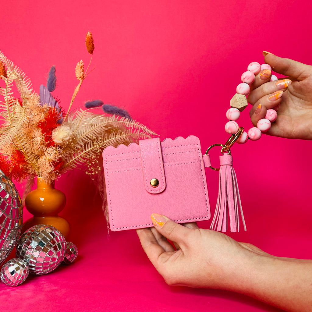 Closeup of woman's hands holding a pink faux leather wallet featuring two back card pockets, a snap closure, attached bracelet made from pink silicone beads, and a pink faux leather tassel. Pink backdrop with an orange vase filled with dried florals sitting next to small disco balls.