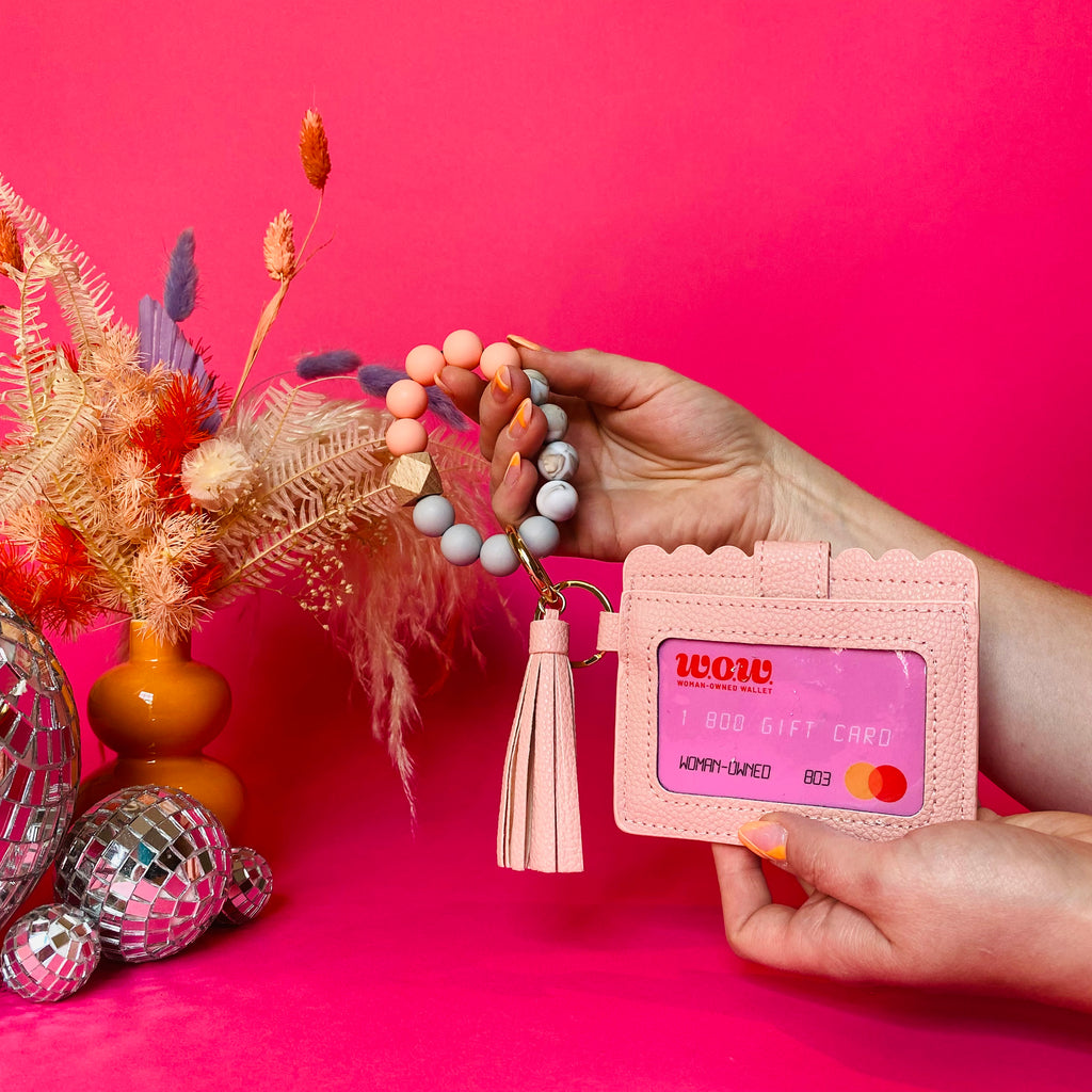Closeup of woman's hands holding a peach faux leather wallet featuring a clear ID pocket, attached bracelet made from peach and grey silicone beads, and a peach faux leather tassel. There is a pink gift card from Woman-Owned Wallet in the ID pocket. Pink backdrop with an orange vase filled with dried florals sitting next to small disco balls.