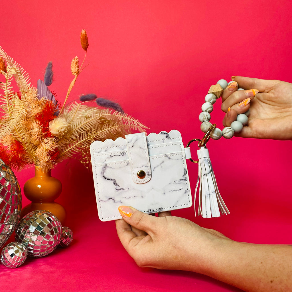 Closeup of woman's hands holding a white faux leather wallet featuring two back card pockets, a snap closure, attached bracelet made from grey and tan silicone beads, and a white faux leather tassel. Pink backdrop with an orange vase filled with dried florals sitting next to small disco balls.