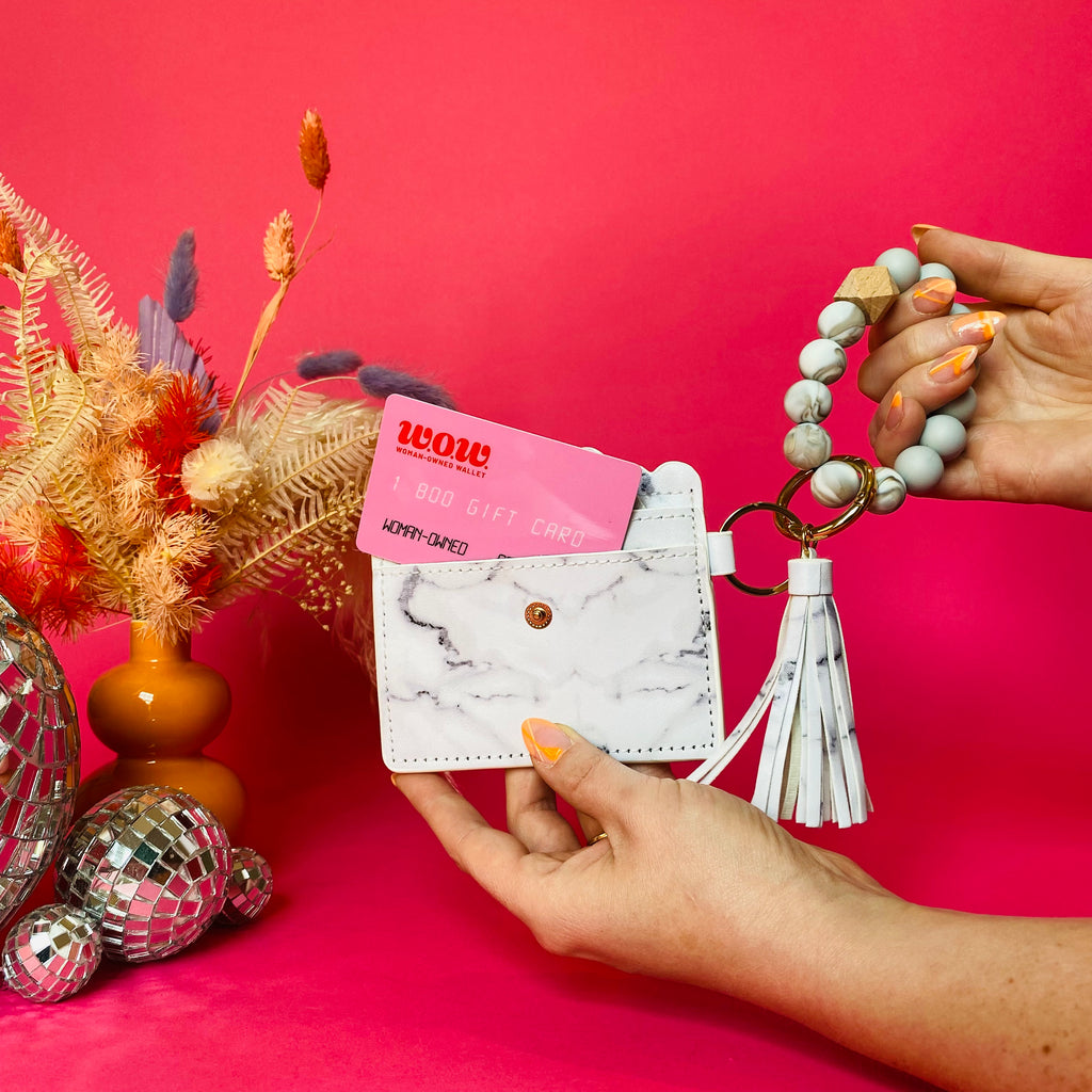 Closeup of woman's hands holding a white faux leather wallet featuring two back card pockets, a snap closure, attached bracelet made from grey and tan silicone beads, and a white faux leather tassel. There is a pink gift card from Woman-Owned Wallet in the ID pocket. Pink backdrop with an orange vase filled with dried florals sitting next to small disco balls.