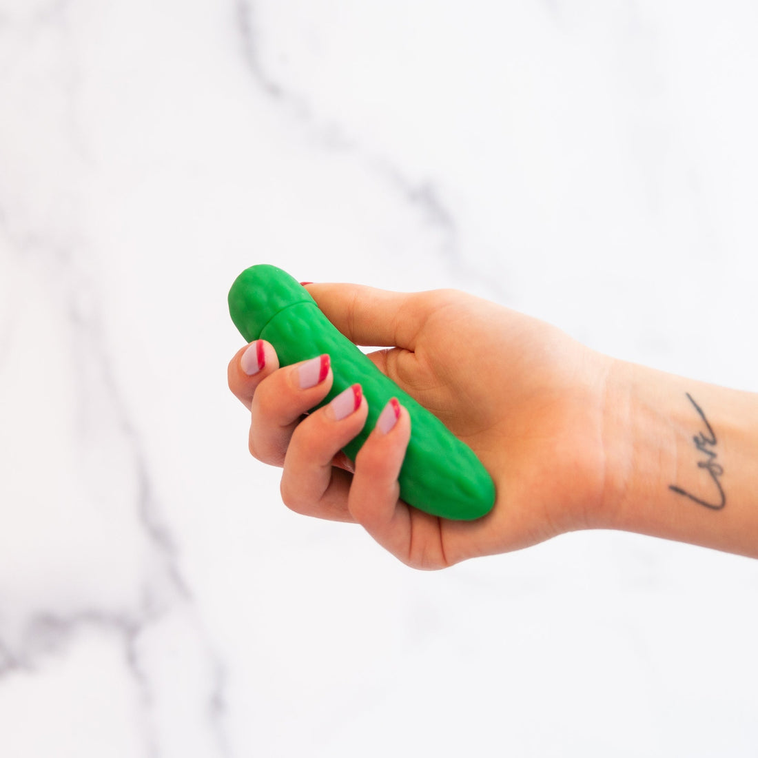 vibrator shaped like a pickle held in a hand with a tattoo that says love on it in front of a white background