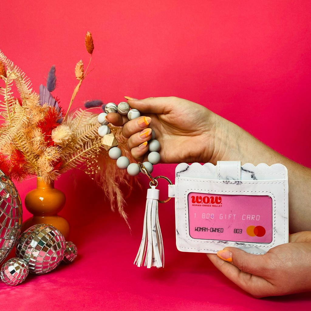 Closeup of woman's hands holding a white faux leather wallet featuring a clear ID pocket, attached bracelet made from grey and white silicone beads, and a tan faux leather tassel. There is a pink gift card from Woman-Owned Wallet in the ID pocket. Pink backdrop with an orange vase filled with dried florals sitting next to small disco balls.
