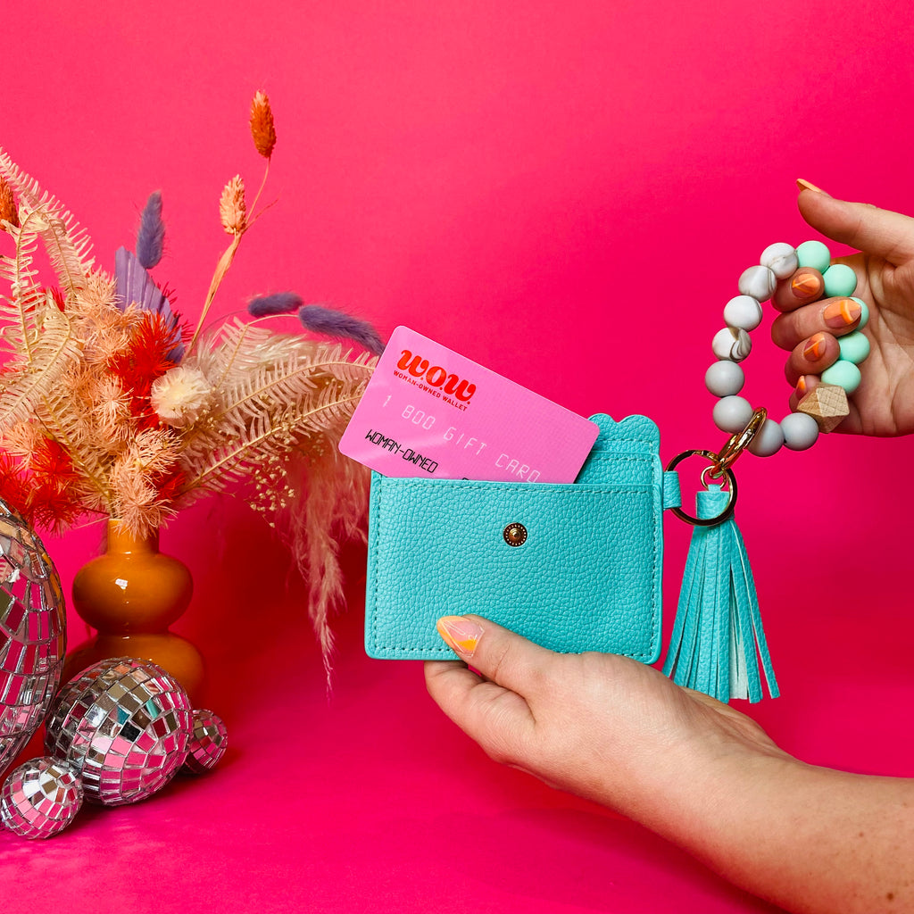 Closeup of woman's hands holding a teal faux leather wallet featuring two back card pockets, a snap closure, attached bracelet made from grey and teal silicone beads, and a teal faux leather tassel. There is a pink gift card from Woman-Owned Wallet in the ID pocket. Pink backdrop with an orange vase filled with dried florals sitting next to small disco balls.