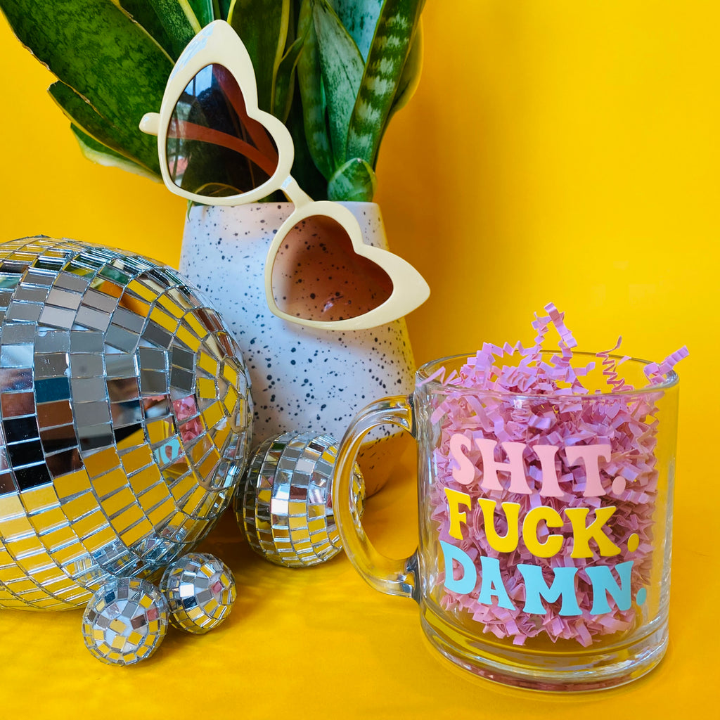 styled shot of plants and disco balls with clear mug with the words "shit fuck damn" in different colors in forefront (with filler in mug)