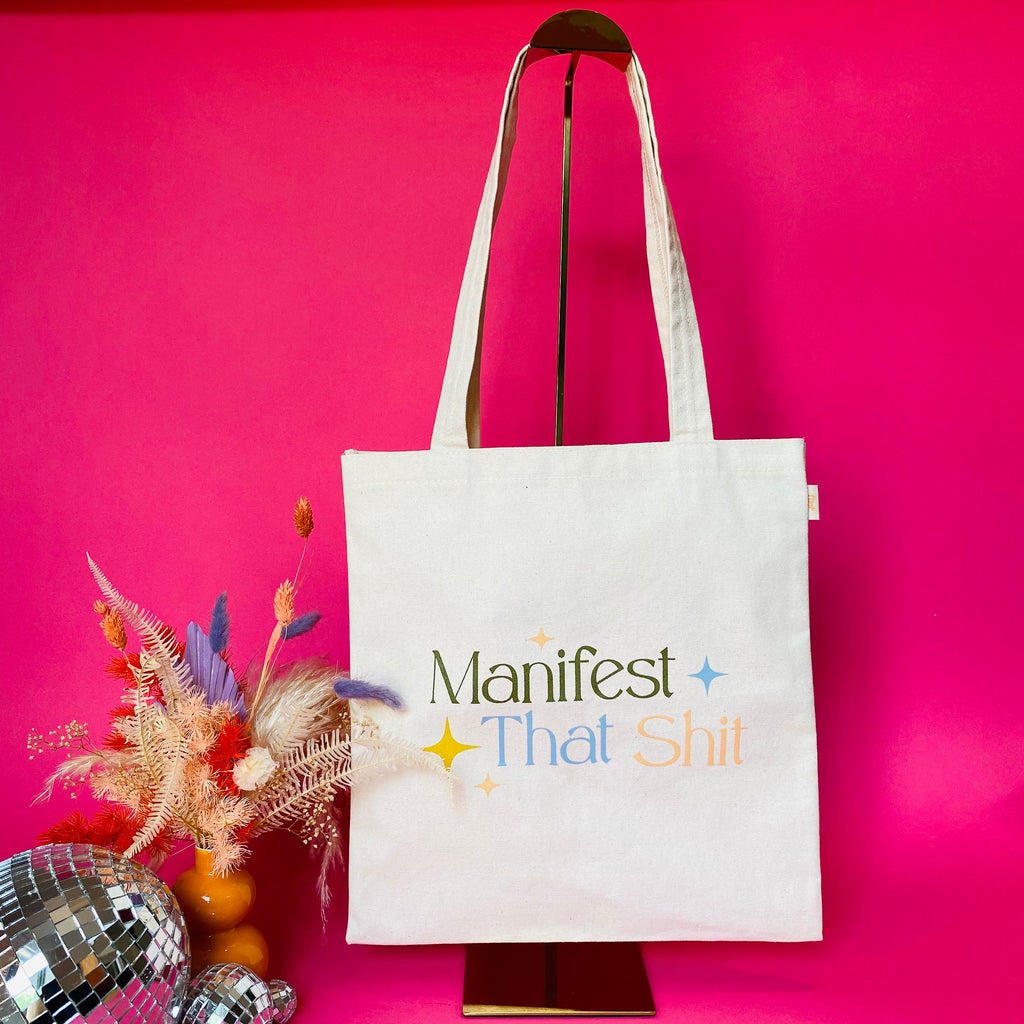 styled shot of Manifest That shit tote against a pink background with dried florals and disco balls
