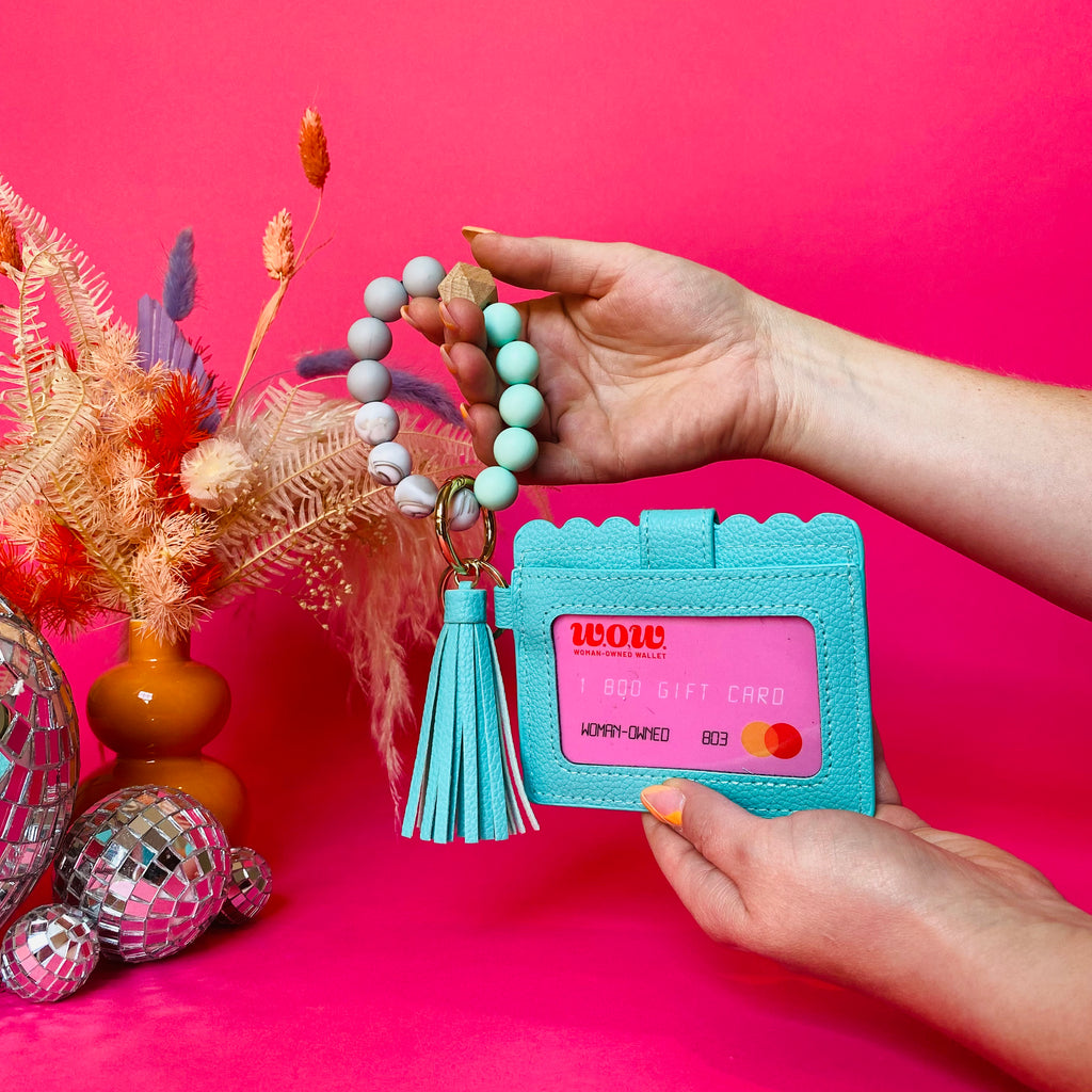 Closeup of woman's hands holding a teal faux leather wallet featuring a clear ID pocket, attached bracelet made from teal and white silicone beads, and a teal faux leather tassel. There is a pink gift card from Woman-Owned Wallet in the ID pocket. Pink backdrop with an orange vase filled with dried florals sitting next to small disco balls.